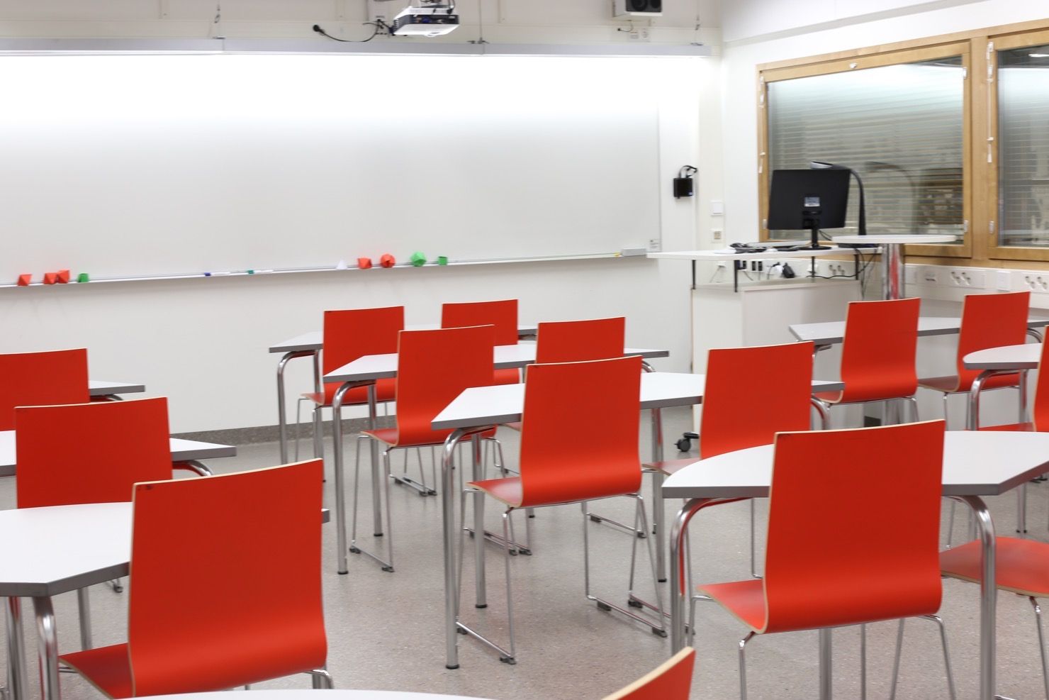Ett klassrumsutrymmen med röda stolar och vita bord. Whiteboard framför stolarna.